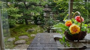 窓際のアンティークテーブルと籠に飾られた大輪のクレイローズ３輪。背景に和庭園。